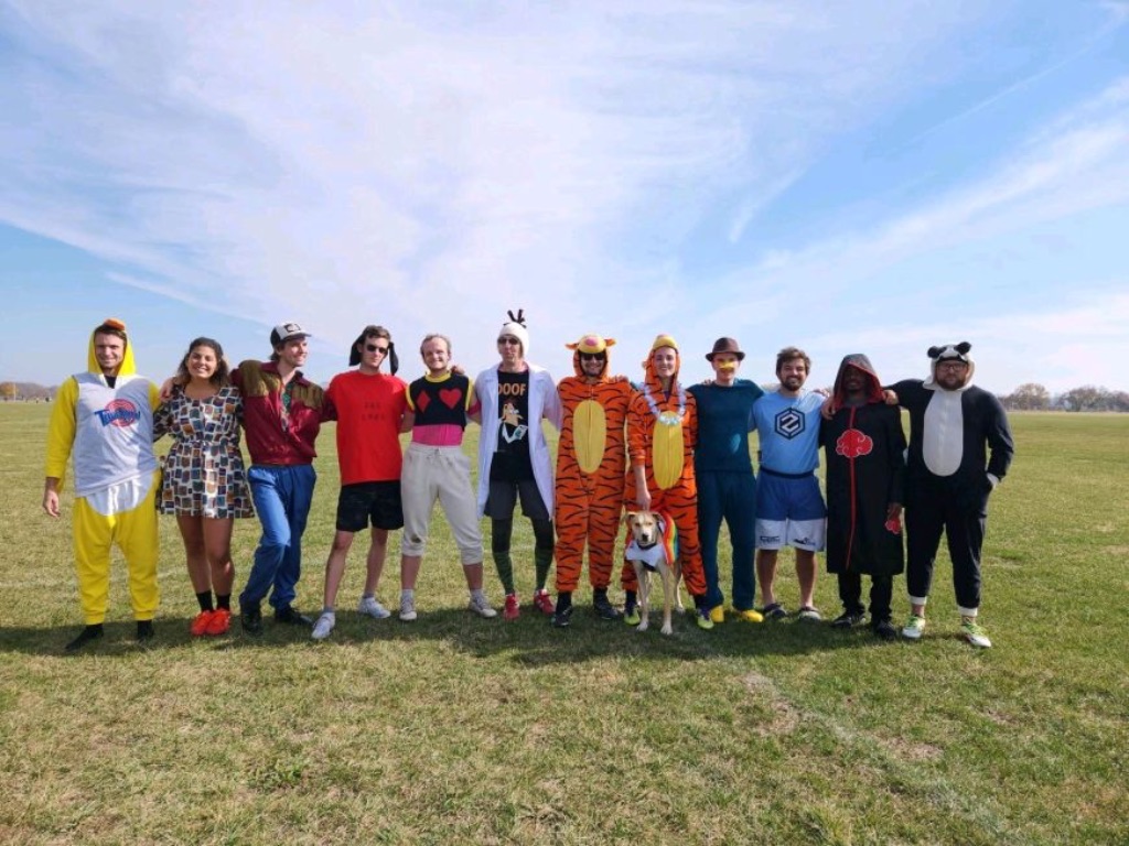 A group of people stand on a grassy field with a huge blue sky behind them. They are all wearing costumes. 