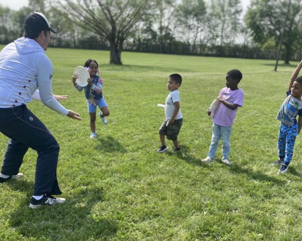 A man in black pants and a white long sleeve shirt stands on a grassy field with four young kids holding discs and smiling. 