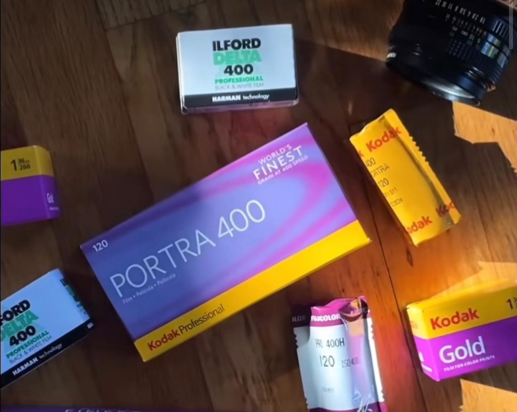 A close up shot of boxes of film on a table. The boxes are various colors of purple, yellow, white. There is a camera lens visible in the upper right corner.