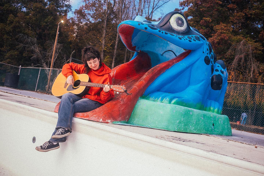 a person sitting on a large blue frog sculpture and playing the guitar. The person’s face is blurred to protect their privacy. The frog sculpture is blue with green and red accents. It has large eyes and a wide open mouth. The person is wearing a red sweatshirt and blue jeans. They are holding a guitar and appear to be playing it. The background is a park or playground with trees and a fence. The photo was taken during the day.