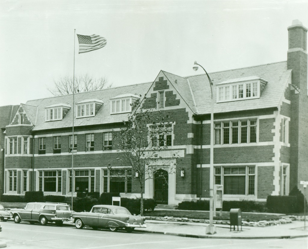 An old black and white picture of a two story brick building. There are two cars parked in front and an American flag is waving at the top of the building.