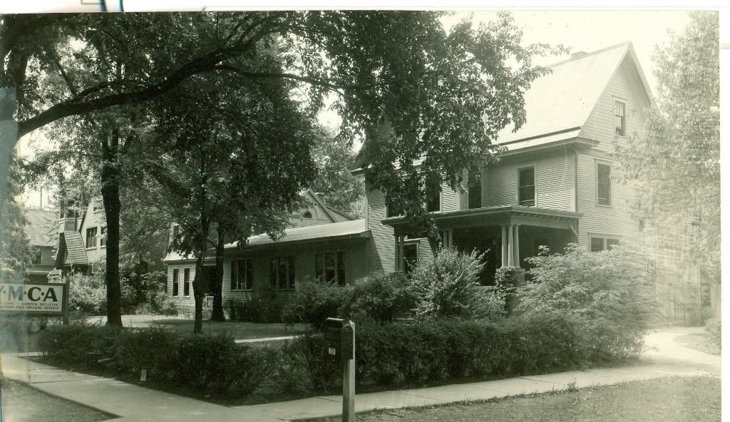 An old black and white picture of a two story brick building. The building is surrounded by hedges and large trees. 