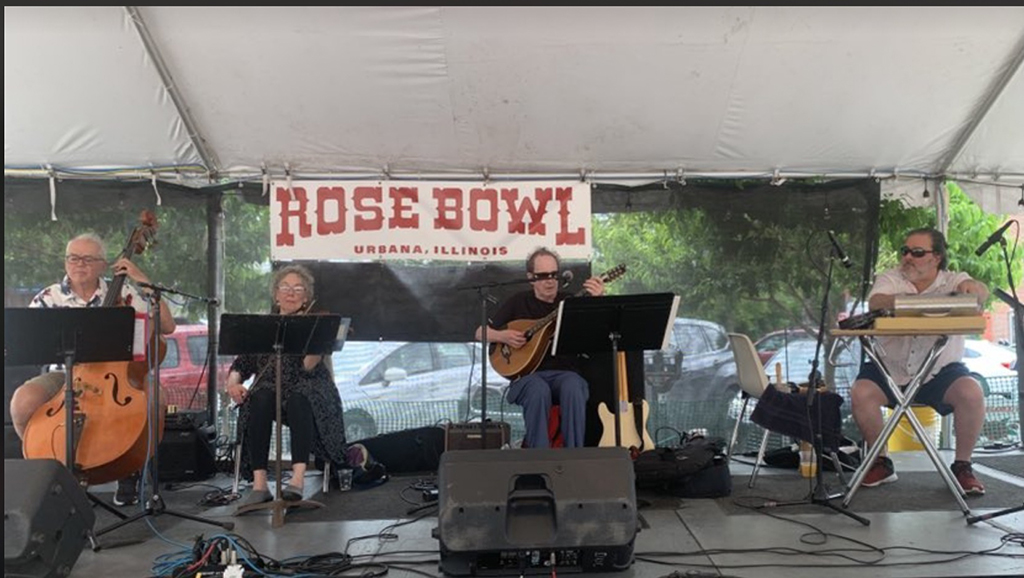 A band is performing on a stage under a tent. The stage has a banner that reads “ROSE BOWL URBANA, ILLINOIS”. The band members are playing various instruments such as a guitar, a keyboard, and a cello. The foreground consists of a speaker.