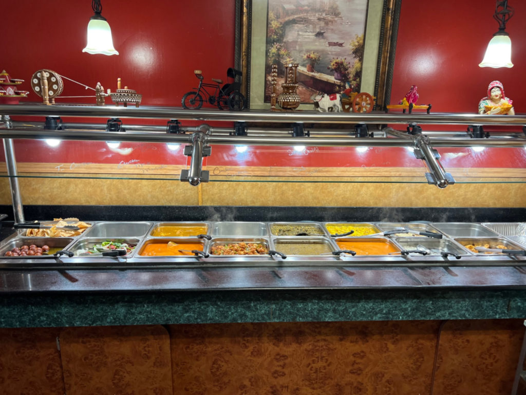 A photo of the hot lunch buffet at Ambar India Restaurant. The wall behind it is bright red, and above the buffet are little trickets and decorative items.