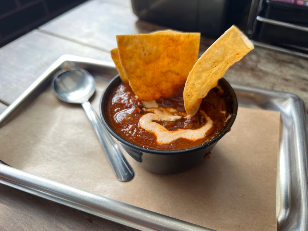 On a metal tray at Black Dog restaurant in Champaign, there is a black cup of dark red sauce with a swirl of chipotle sour cream and three tortilla chips dusted with sauce. The brisket chili is included in a fall flavors list.