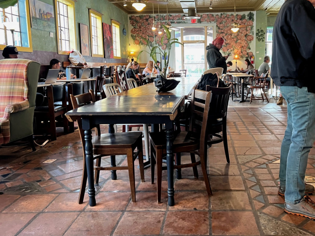 Inside Caffe Paradiso, there are various patrons each working solo inside the coffee shop. Mismatched wooden chairs are tucked into mismatched tables. Art and murals cover the walls.