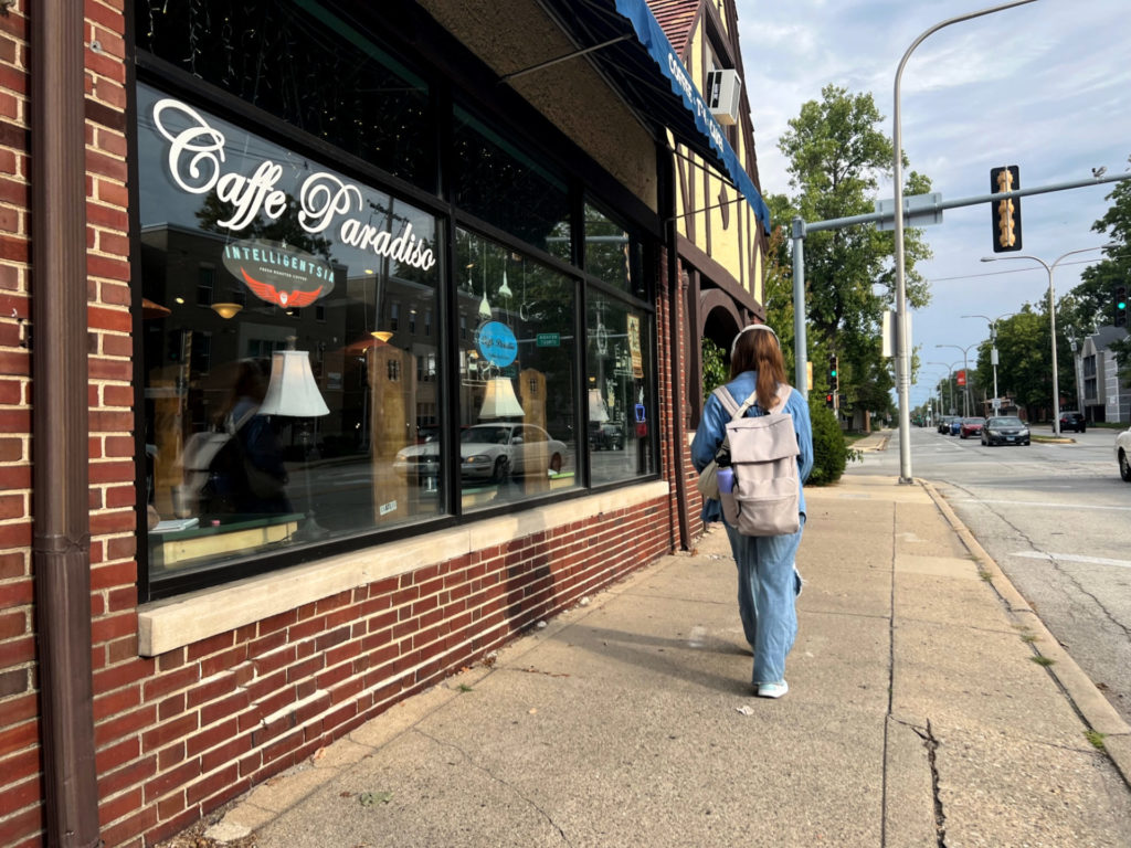 The exterior of Caffe Paradiso on Lincoln Avenue has big windows on a brick building. A student with a pink backpack and headphones walks along the sidewalk as cars drive down Lincoln Avenue.
