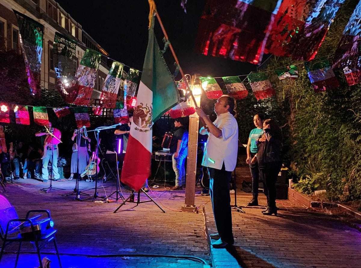 A man stands on an outdoor patio holding a Mexican flag for a ceremony, while a band plays off to the side.