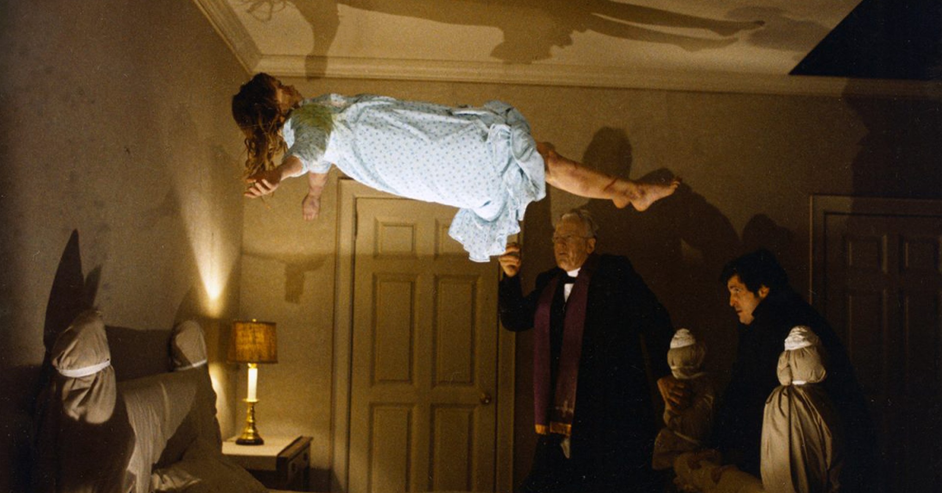 A screenshot from The Exorcist. A young girl in a nightgown is floating above her bed, with adults surrounding her.
