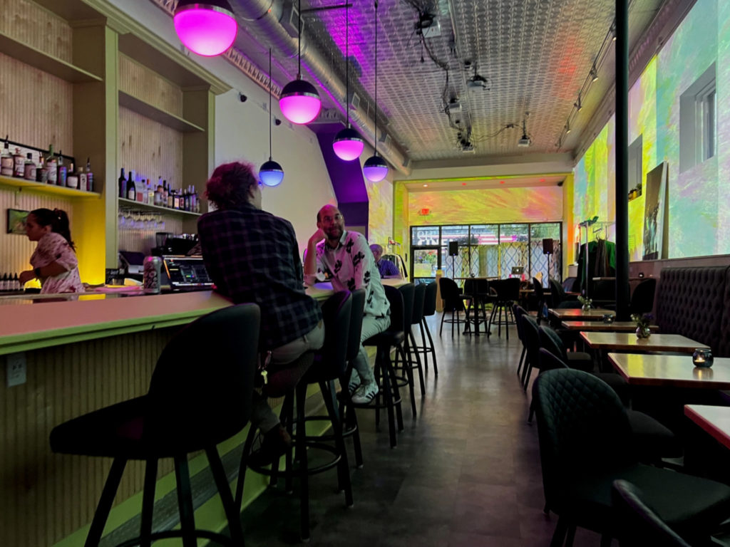 The interior of Gallery Art Bar has low tables on the right and bar seating on the left. Pendant lights are in shades of pink and purple.