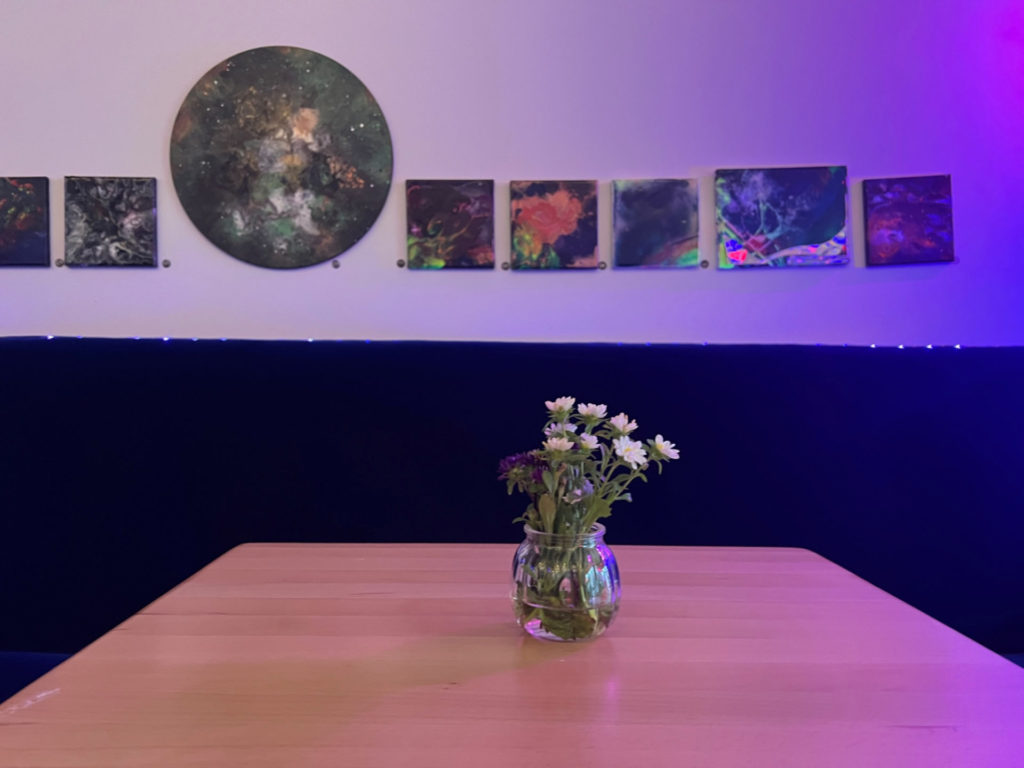 On a table, there is a clear vase with fresh cut flowers. There is a blue velvet couch with art hanging above it.