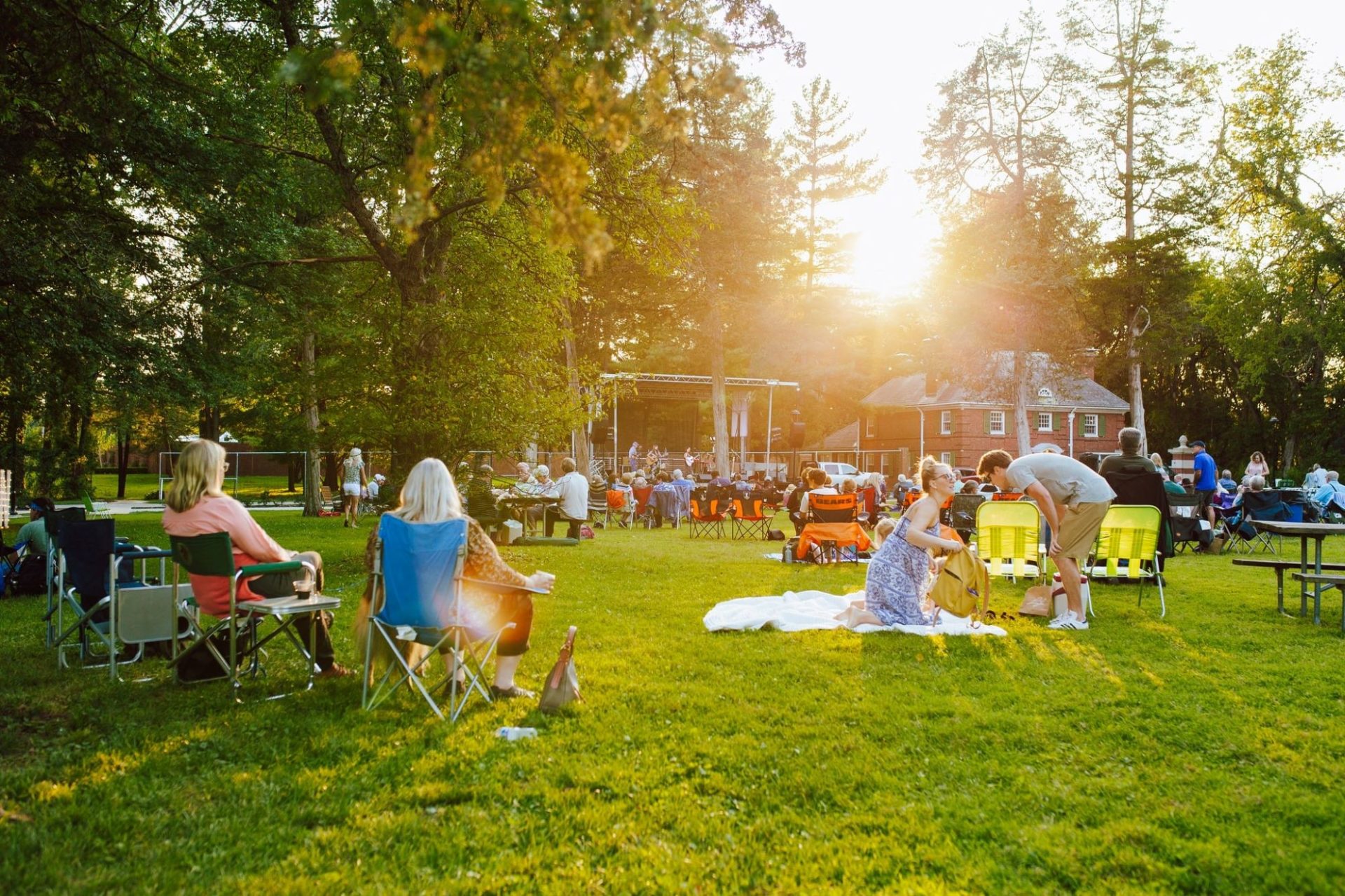 An outdoor concert with people gathered on a grassy lawn on blankets and in chairs. The sun is setting behind the trees.