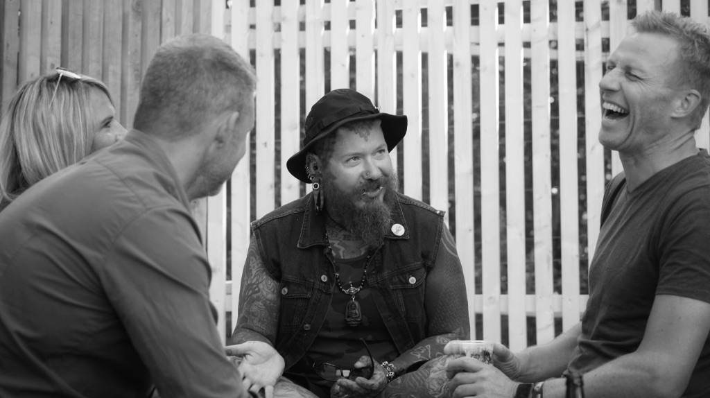 Three white men sitting in a triangle talking to each other and laughing and smiling. The man in the middle wears a black vest and hat and has many piercings while the other two  men wear t-shirts. 