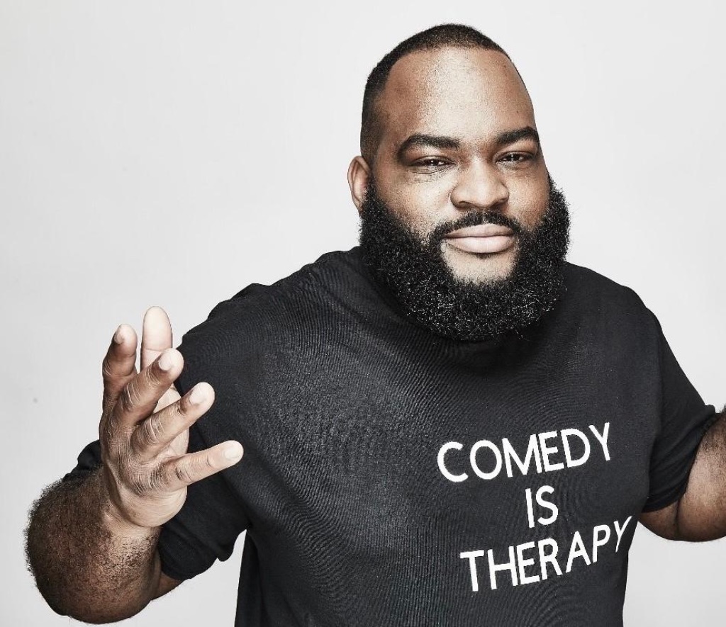 A black man wearing a black t-shirt with white print that says "comedy is therapy". He has his arms raised in a shrug position. He has close cropped hair and a full beard and mustache. 