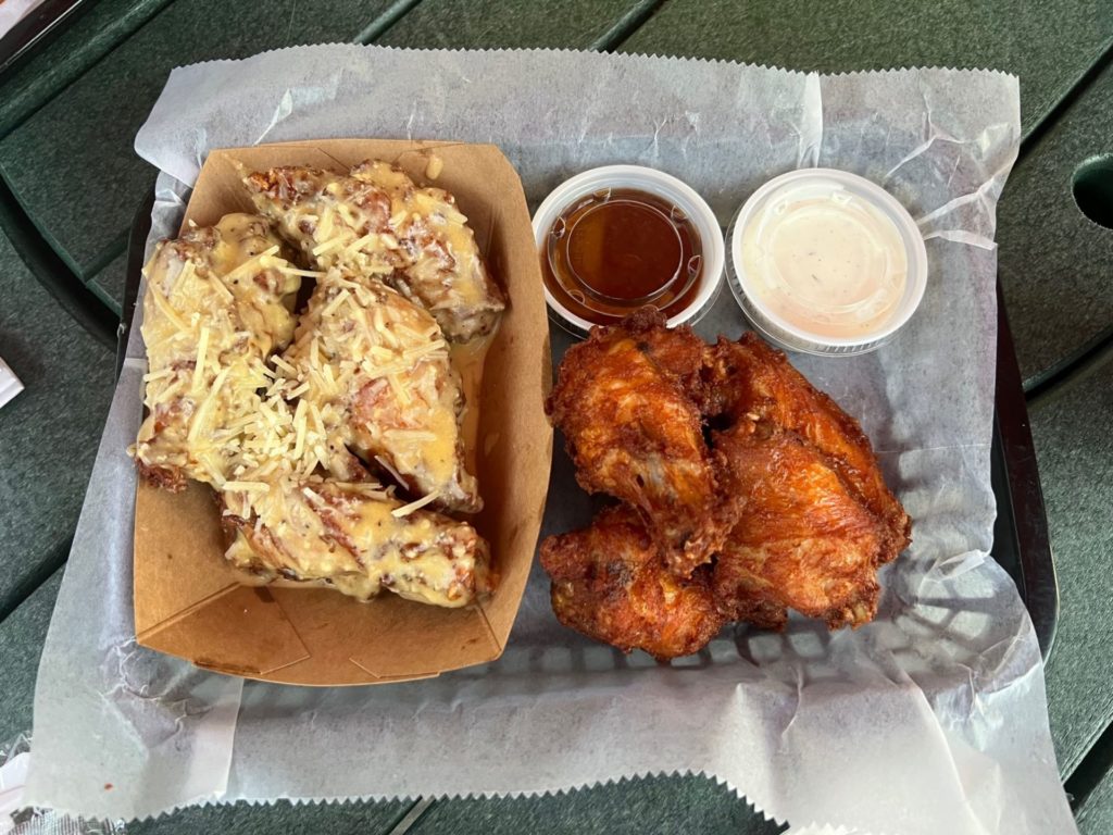 A basket lined with wax paper. On one side is a smaller container with garlic parmesan wings, and on the other is a pile of wings and two dipping sauces.