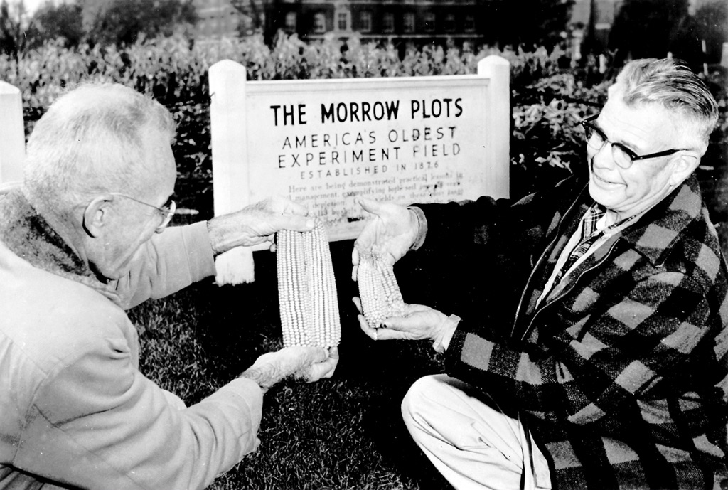 A black and white picture of two men jkneeling in a field comparing ears of corn. Behind them are planted rows of corn and a white sign that says The Morrow Fields.