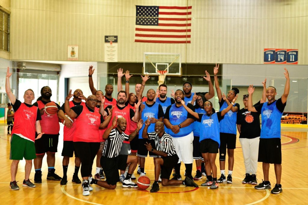 A group of people are gathered on a basketball court. Several are wearing red jerseys, and several are wearing blue jerseys. There are two referees in black and white shirts kneeling in front.