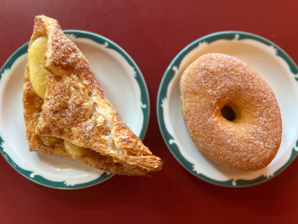 Apple turnover on a white plate with a green border and a cinnamon sugar doughnut from Rick's Bakery on the same style plate beside it.