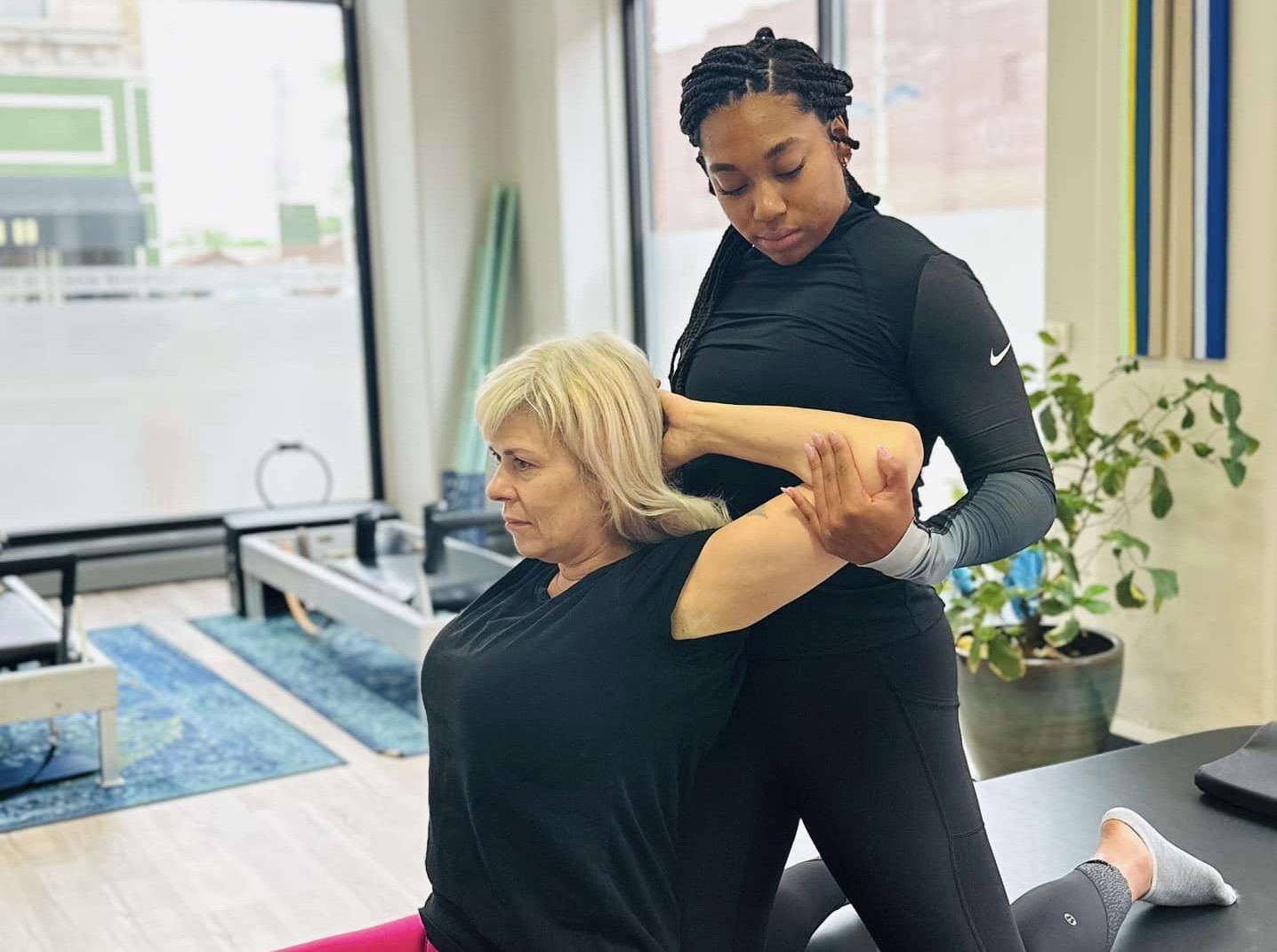 A white woman is sitting on a bench, hands behind her head, with elbows out. A Black woman therapist is positioned behind her, stretching her arms.