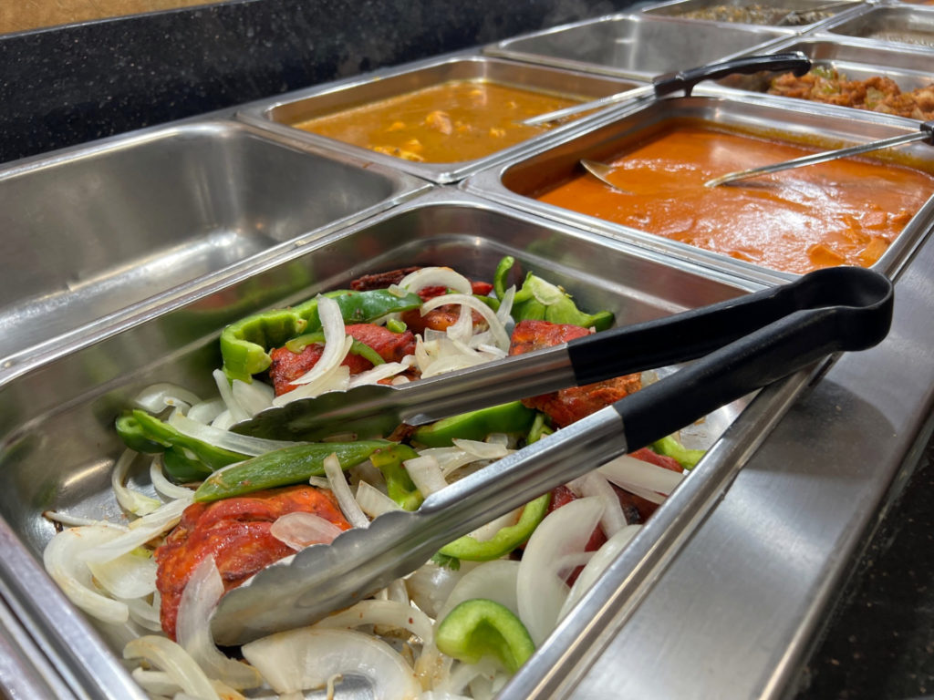 The tandoori chicken on the buffet line has a silver and black pair of tongs over mostly raw white onion and green bell pepper slices.