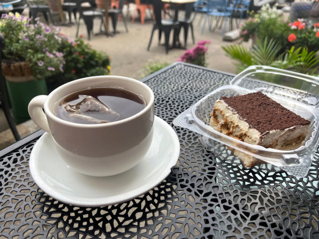 A mug of tea on a saucer with a small chip and a to-go slice of tiramisu in a clear, plastic clamshell container.