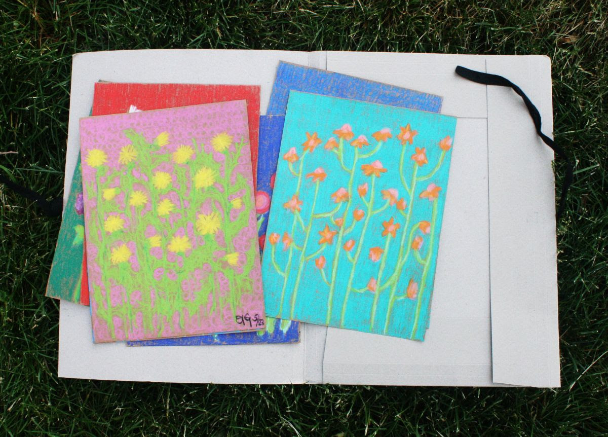 Works by Anna Gutsch lay on a white cardboard in the grass. They are brightly colored simple floral drawings