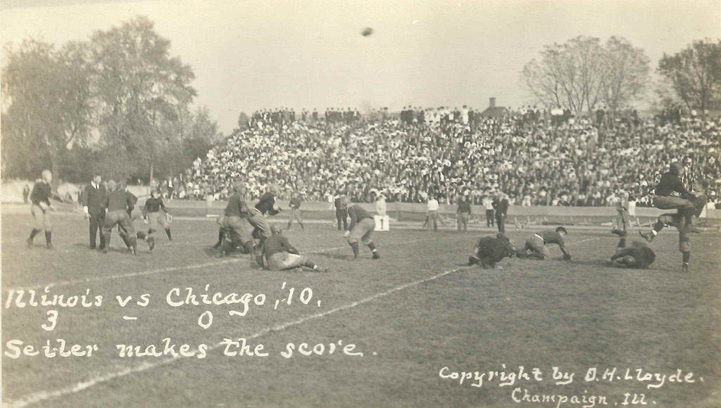 A very old black and white photo of the fist homecoming football game from the year 1910. There is are two teams playing football while a crowd watches from bleachers in the distance. 
