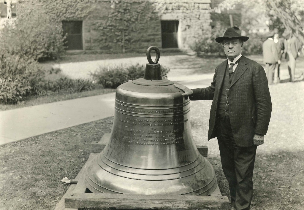 A black and white picture of a man in a suit and hat standing next to a giant bell that comes up to his shoulder