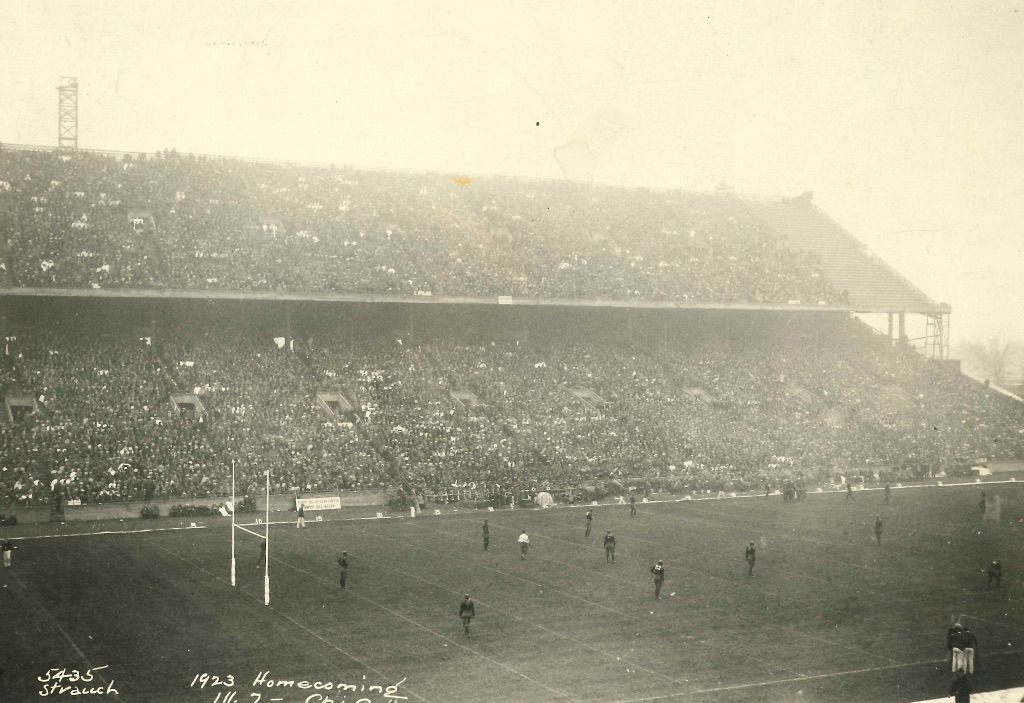 A very old picture of the u of I memorial stadium overlooking players on the filed and a seats full of people watching the game.