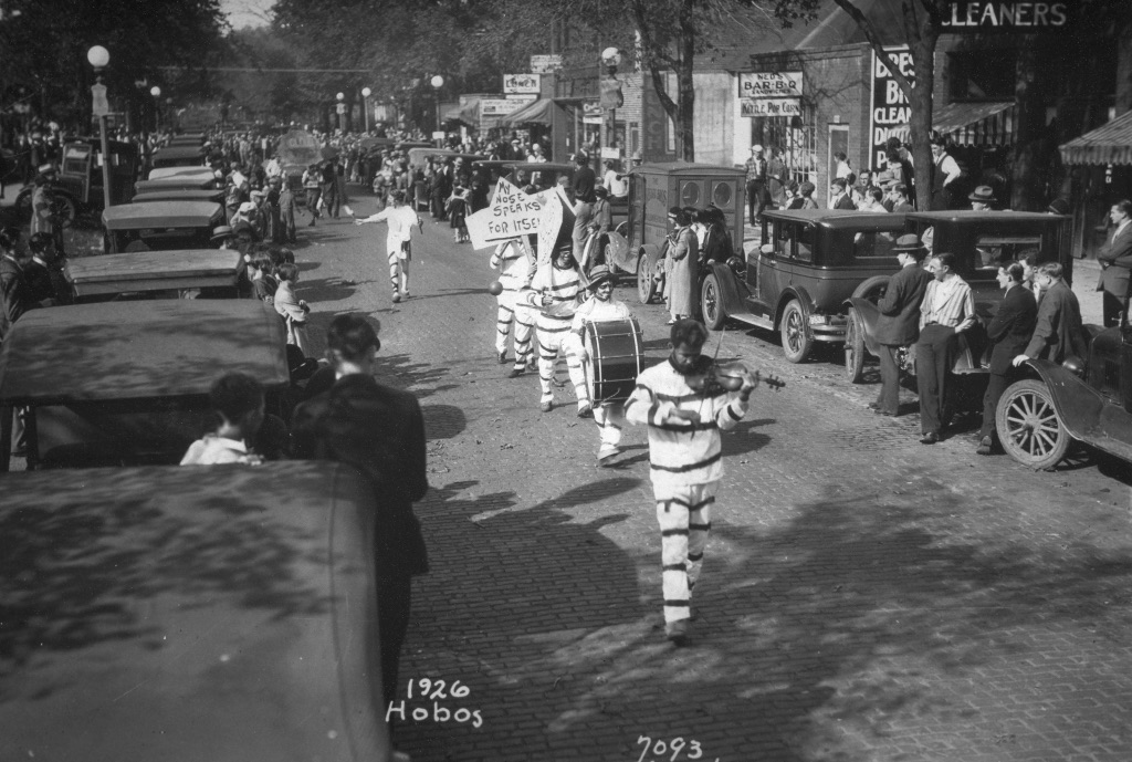 a black and white picture of men walking down the street in black and white striped outfits playing instruments including a violin. While people watch from the side of the street. 