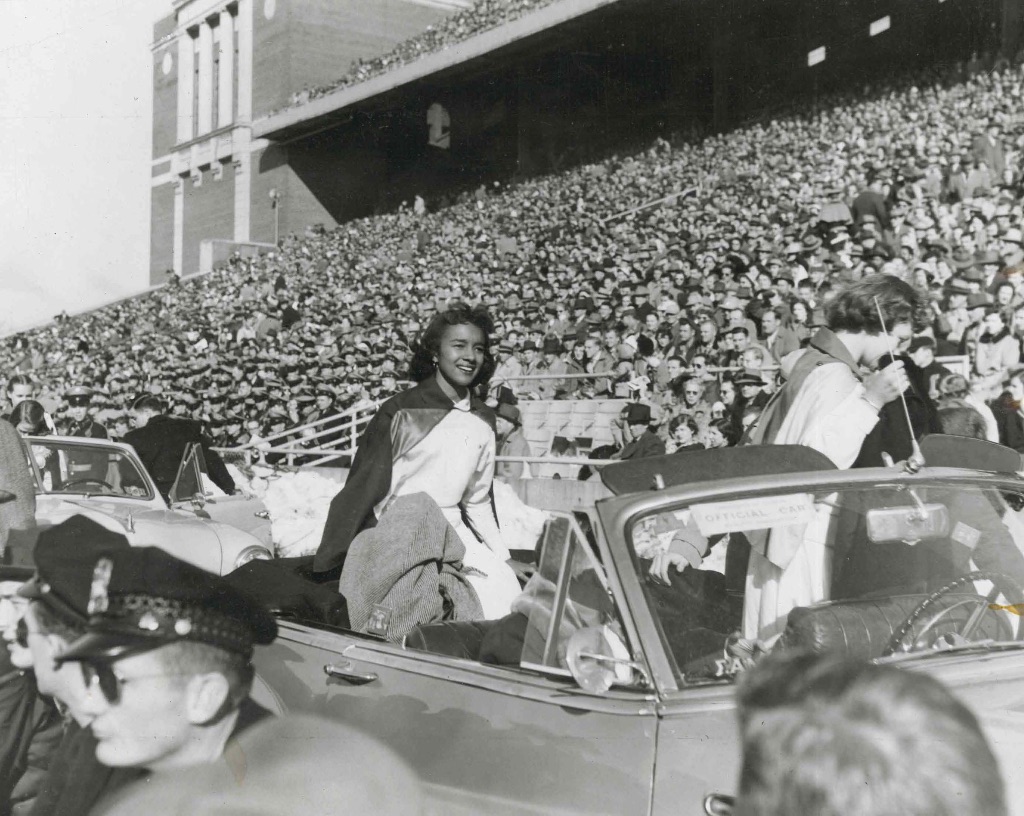 A black woman sits on the back of an open air convertible in a white dress driving on the filed through the stadium. 