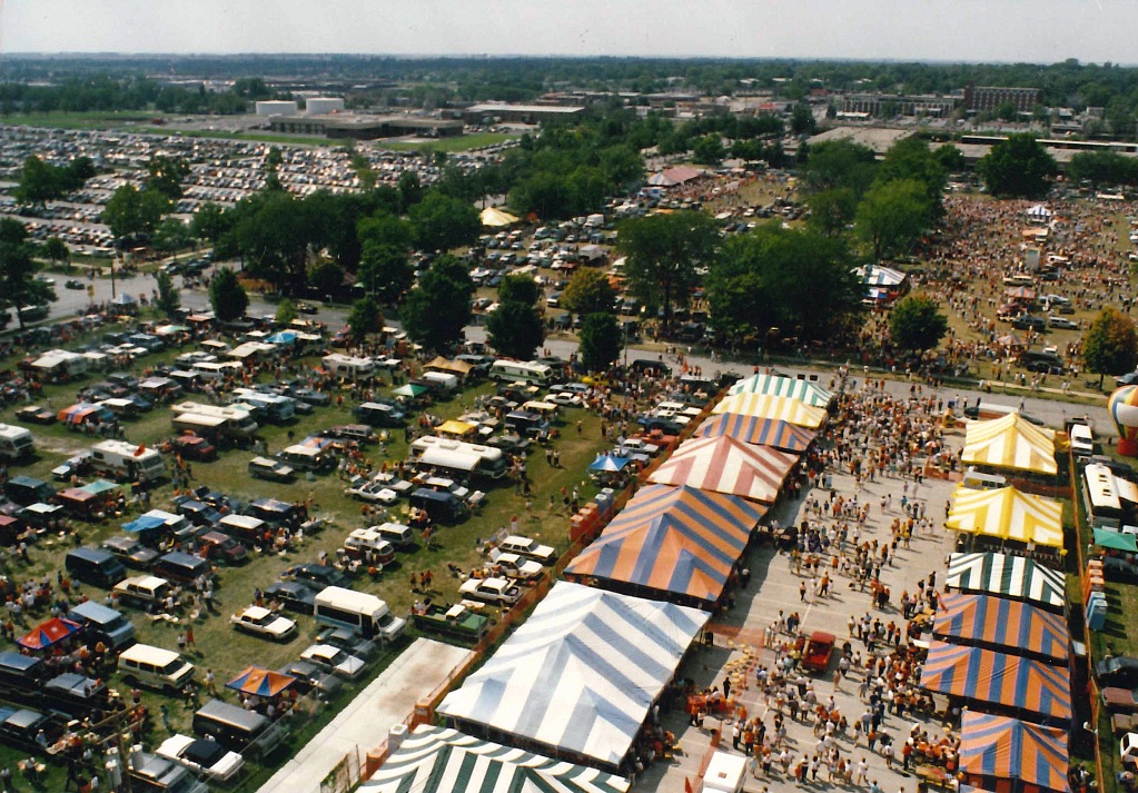 An aerial shot of a tailgate lot with rows of cars and colorful striped tents in a big field.
