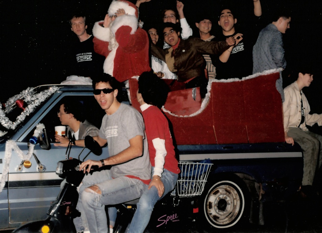 A group of people ride on a car and a moped next to it. They are wearing sunglasses and one person is dressed as Santa clause. 