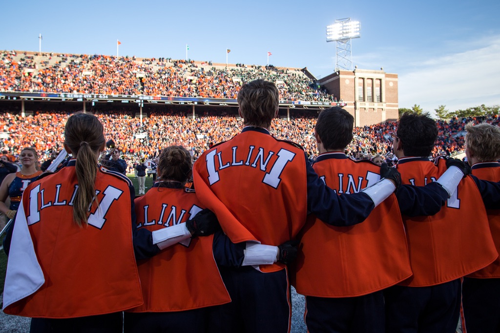 Several University of Illinois Marching Band students pictured from behind with their arms around each other. They are standing on a football field. They are wearing bright orange coats that say "illini" on the back.