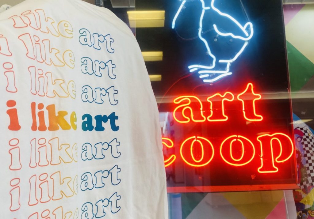 a rainbow colored t shirt that says "I like art" is displayed in front of the neon art coop sign in their display window