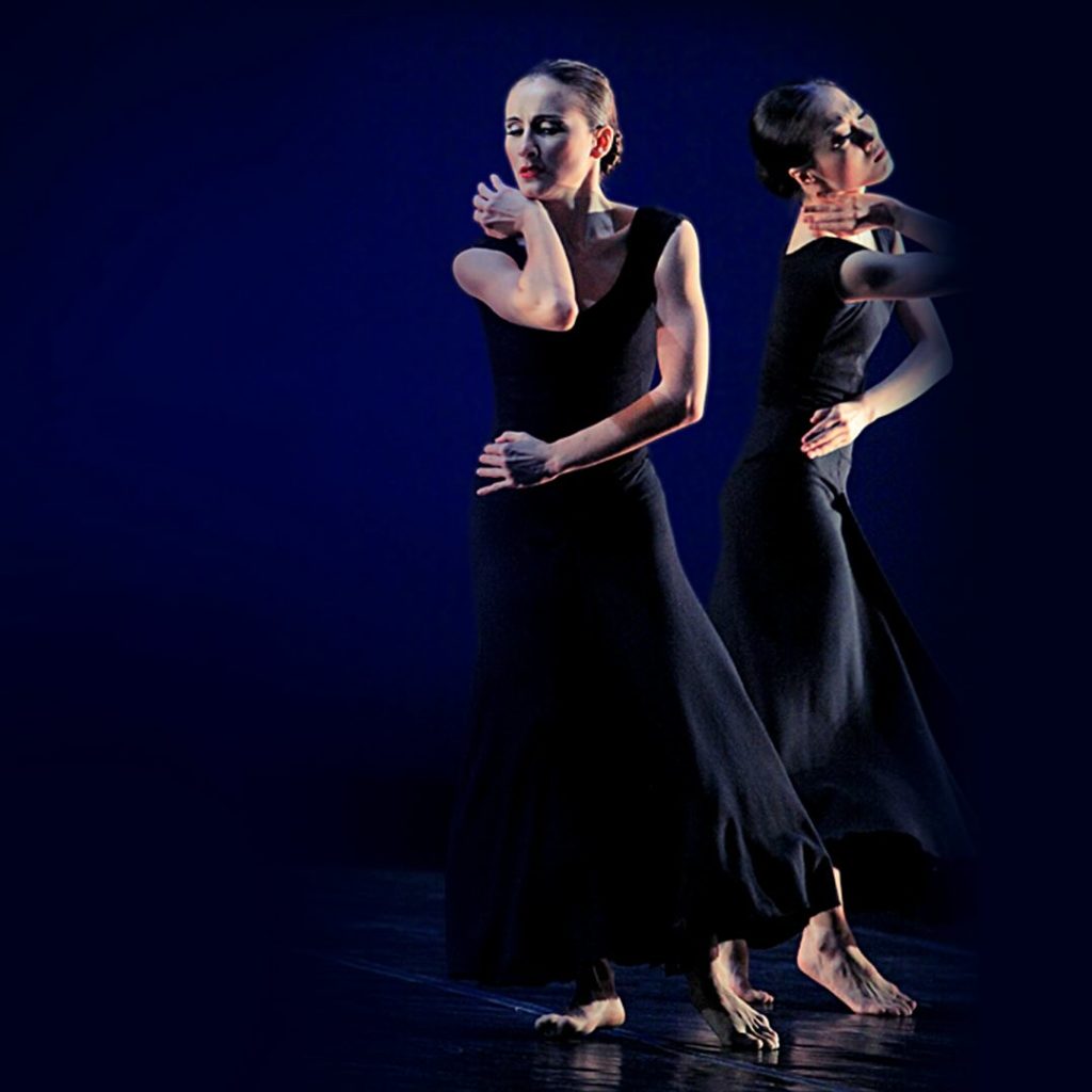 two female dancers wearing floor length black dresses dance on a dimly lit stage. They are mirroring each other