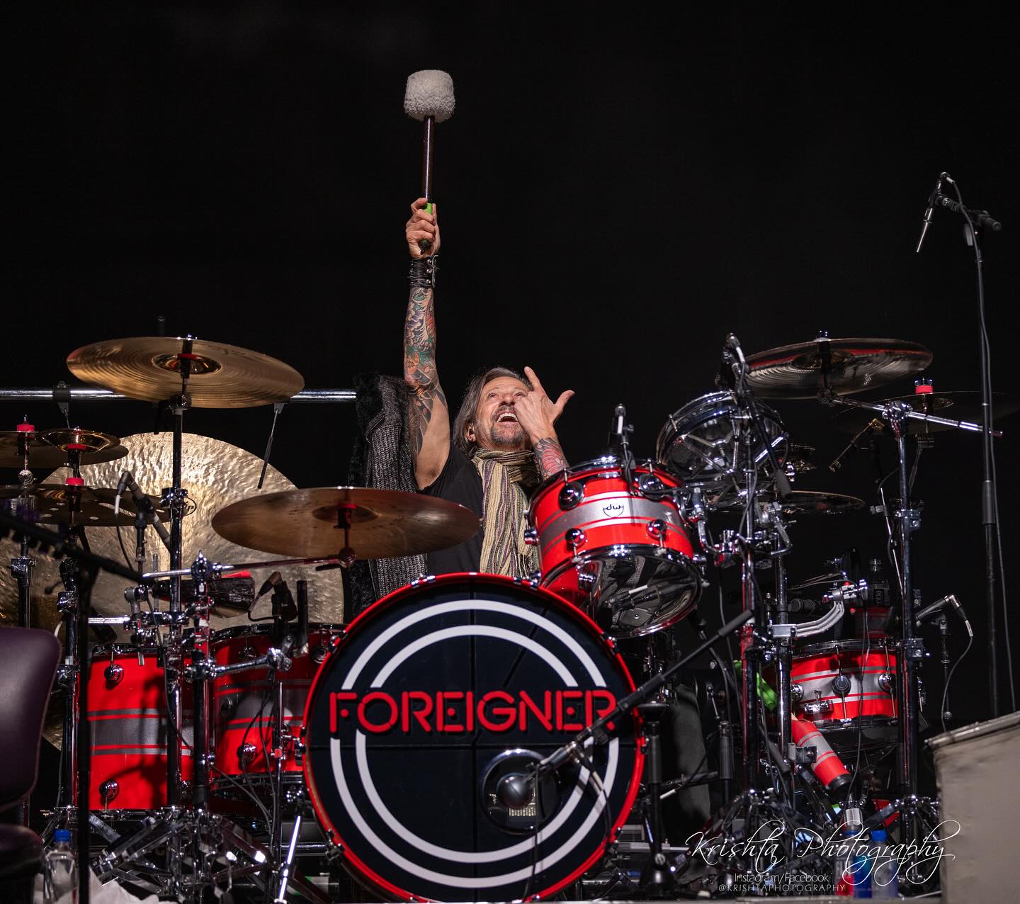 A white man sits behind a red and black drum kit. The kit says "Foreigner." The man has his right arm with a drum mallet lifted. His left hand is pointing to his right hand.