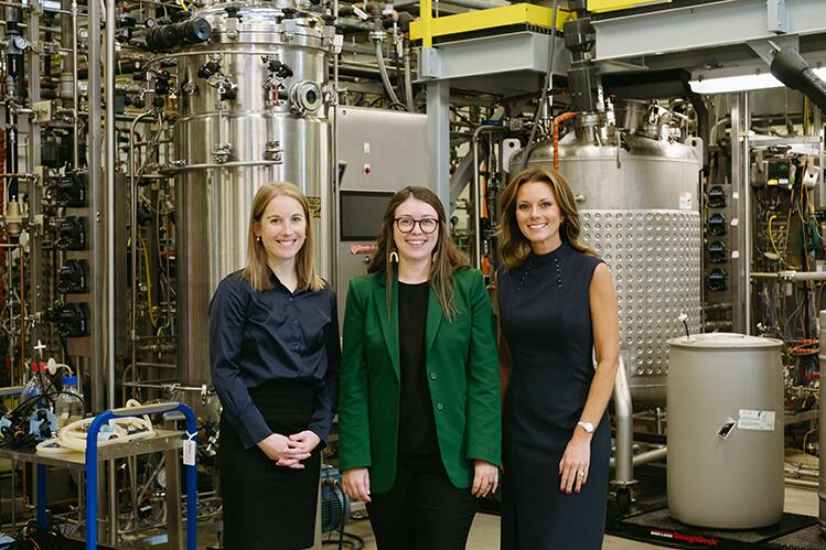Three white women in professional dress, standing in a row in a manufacturing facility.