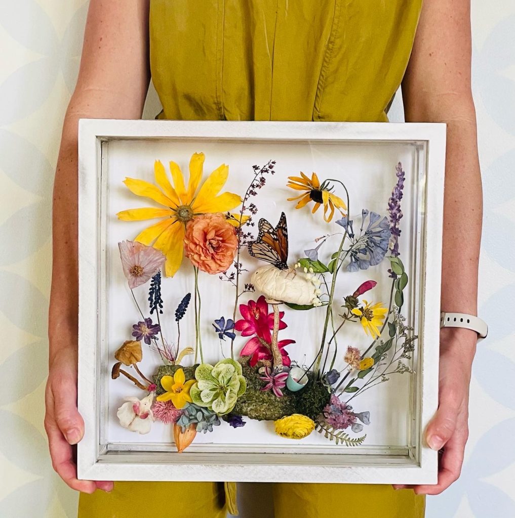 Hughes, with only her torso and arms visible, holds a small white shadowbox filled with flowers