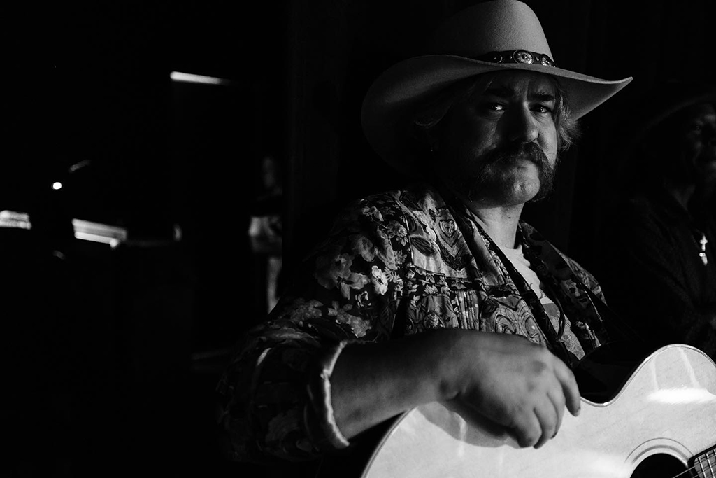 In the image, there’s a person playing a guitar. They are wearing a cowboy hat and a floral shirt. The image is in black and white, and the background is dark and blurred.