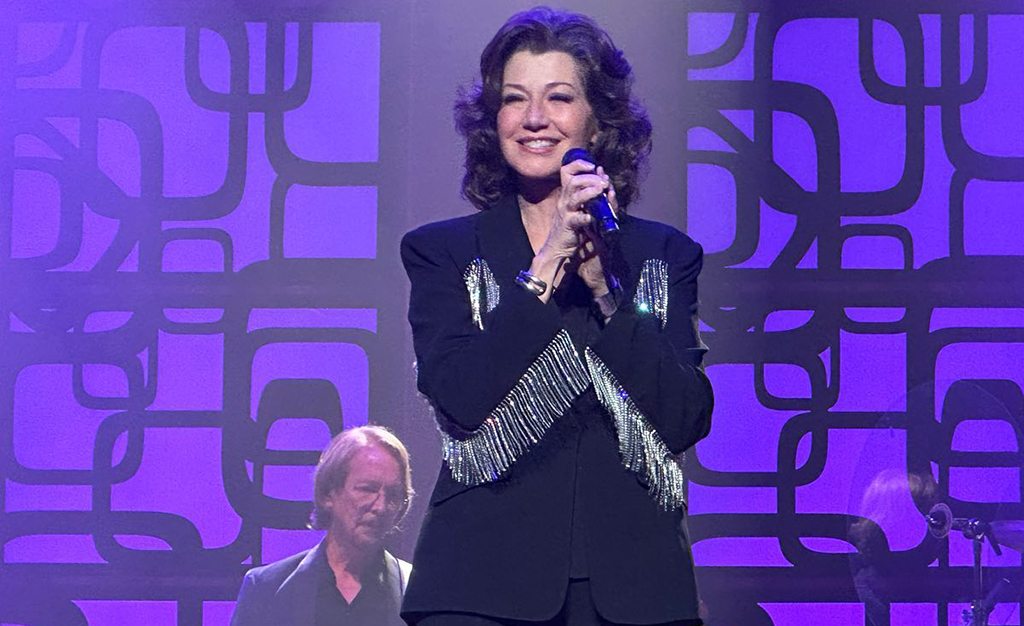 A person is on stage, microphone in hand, dressed in a black suit adorned with silver sequins on the sleeves. Behind them, another individual sits against a backdrop of a purple wall with a geometric pattern.