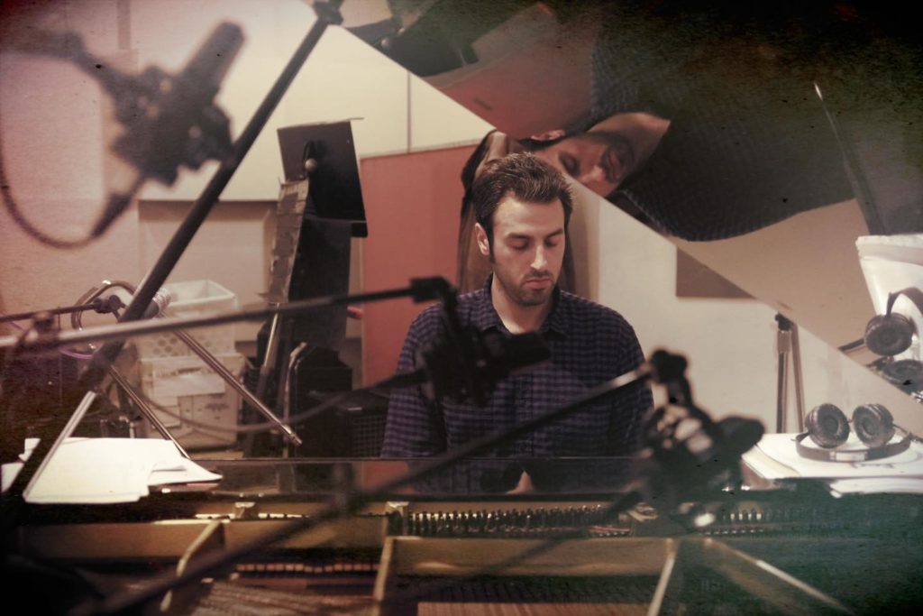 The image presents a recording studio. In the center, a blurred figure can be seen. The room is equipped with a desk featuring a computer monitor, a keyboard, and a microphone. A piano with sheet music rests in the foreground, and several microphone stands are scattered throughout the space. The image has a sepia tone and appears to be taken from a low angle.
