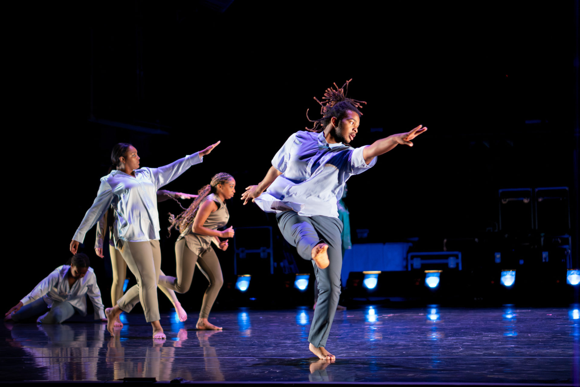 A male Black dancer is front and center with other dancers behind performing a modern dance with arms outstretched. They are all wearing blues and neutrals satin long sleeve tops and leggings.