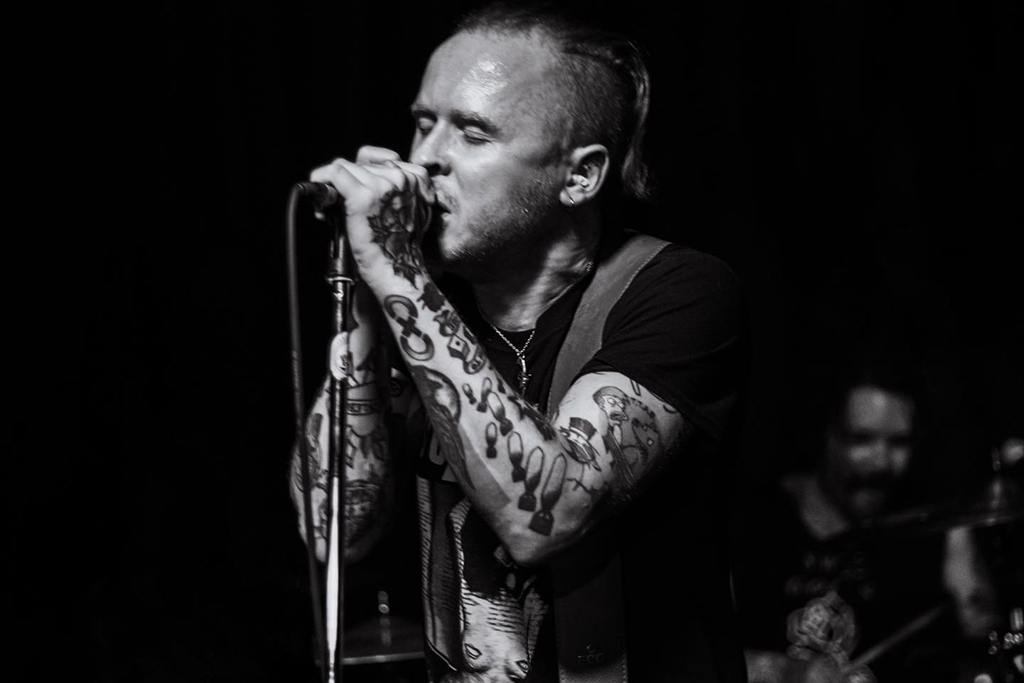 A black and white photo where a person takes center stage, their voice resonating through the microphone they hold. Their attire is a simple black tank top, a canvas for the intricate tattoos that decorate their arms. In the background, another figure is immersed in the rhythm of the drums. Spotlights illuminate the stage, casting an ethereal glow on the performers and setting the mood for the performance.
