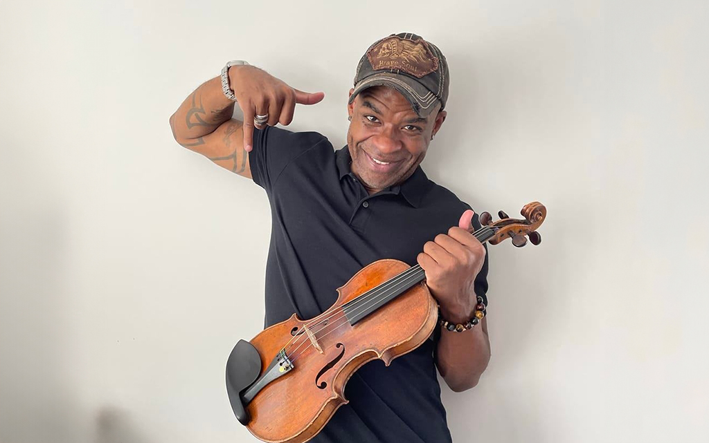 A person is captured in the act of pointing to a violin they hold in their right hand. They are attired in a black polo shirt and a baseball cap. A beaded bracelet adorns their left wrist, which also showcases a series of tattoos. The backdrop is a plain white wall.