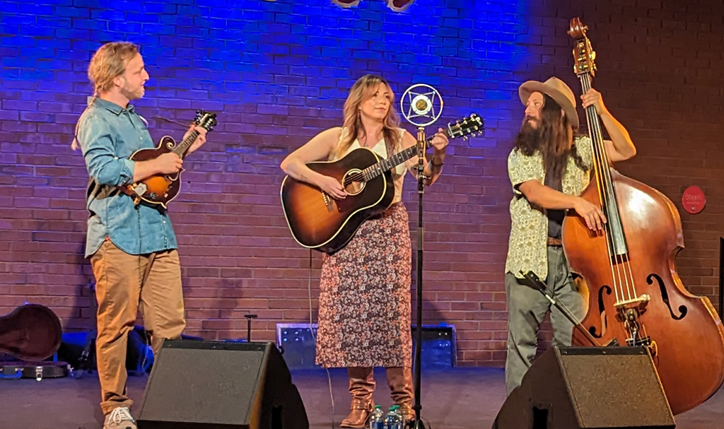 Three musicians performing onstage with a brick wall backdrop. The one on the left plays the mandolin, the middle one is positioned in front a an old-timey microphone holding a guitar, and the one on the right is playing an upright bass.