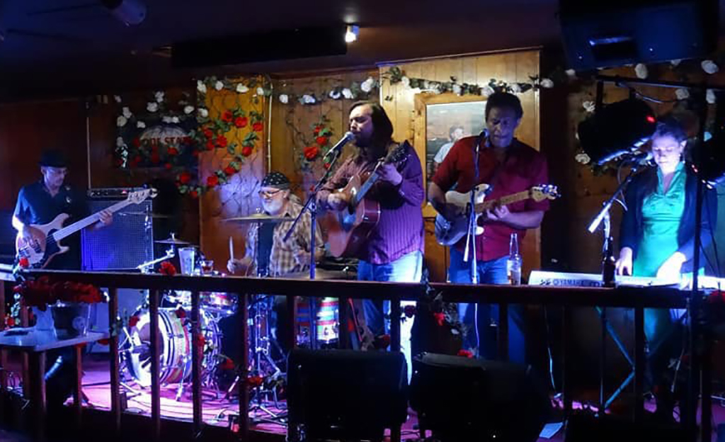 A band is playing on a stage in a bar. There are five people on the stage, all dressed in casual clothing. The person on the left is playing a guitar, the person in the middle is playing a banjo, the person on the right is playing a guitar, and the person on the far right is playing a drum set. The stage is decorated with Christmas lights and flowers.