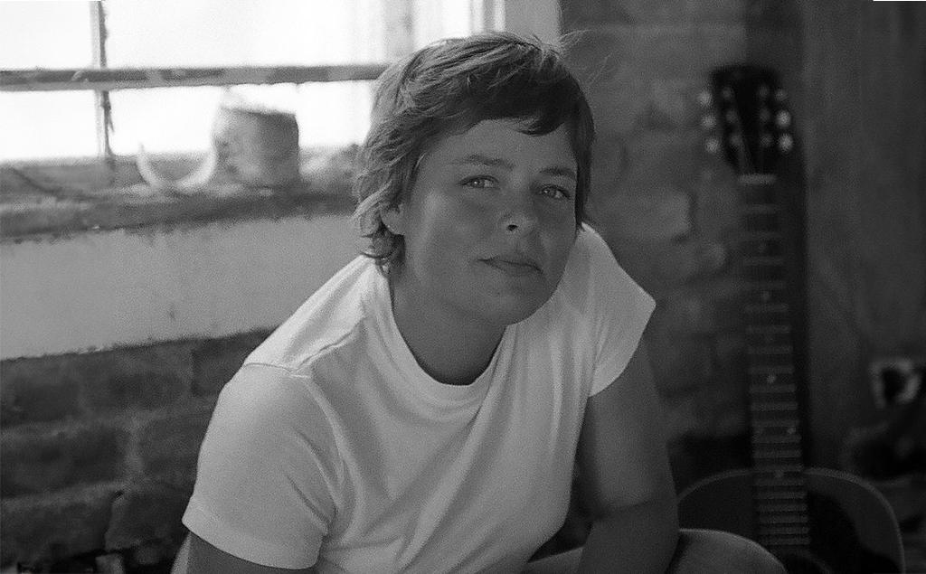 A person is perched on a window sill, their back to the window. They are dressed in a white t-shirt. A guitar leans against a brick wall in the background. The image is in black and white.