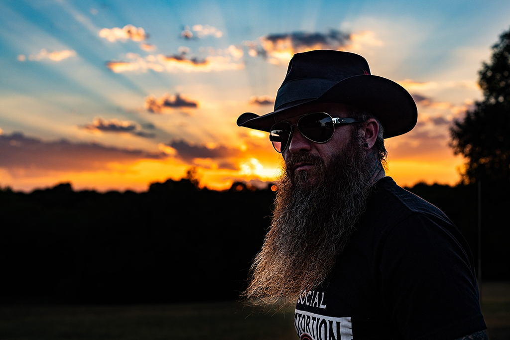 An individual stands against a backdrop of a breathtaking sunset, donned in a black cowboy hat and a black t-shirt with white text. Their long beard adds to their distinctive look as they gaze into the distance.