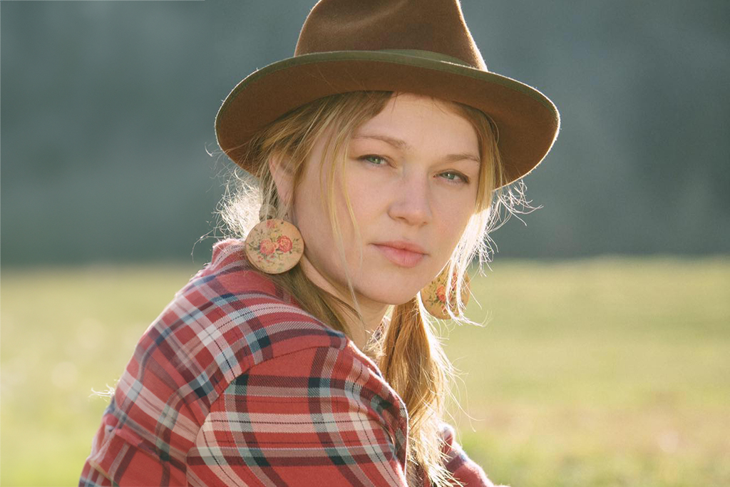 The image captures a person standing in a grassy field with trees in the background. The individual is wearing a brown hat and a red plaid shirt. Large round earrings with a floral design are noticeable. The image has a soft, dreamy quality with warm lighting.